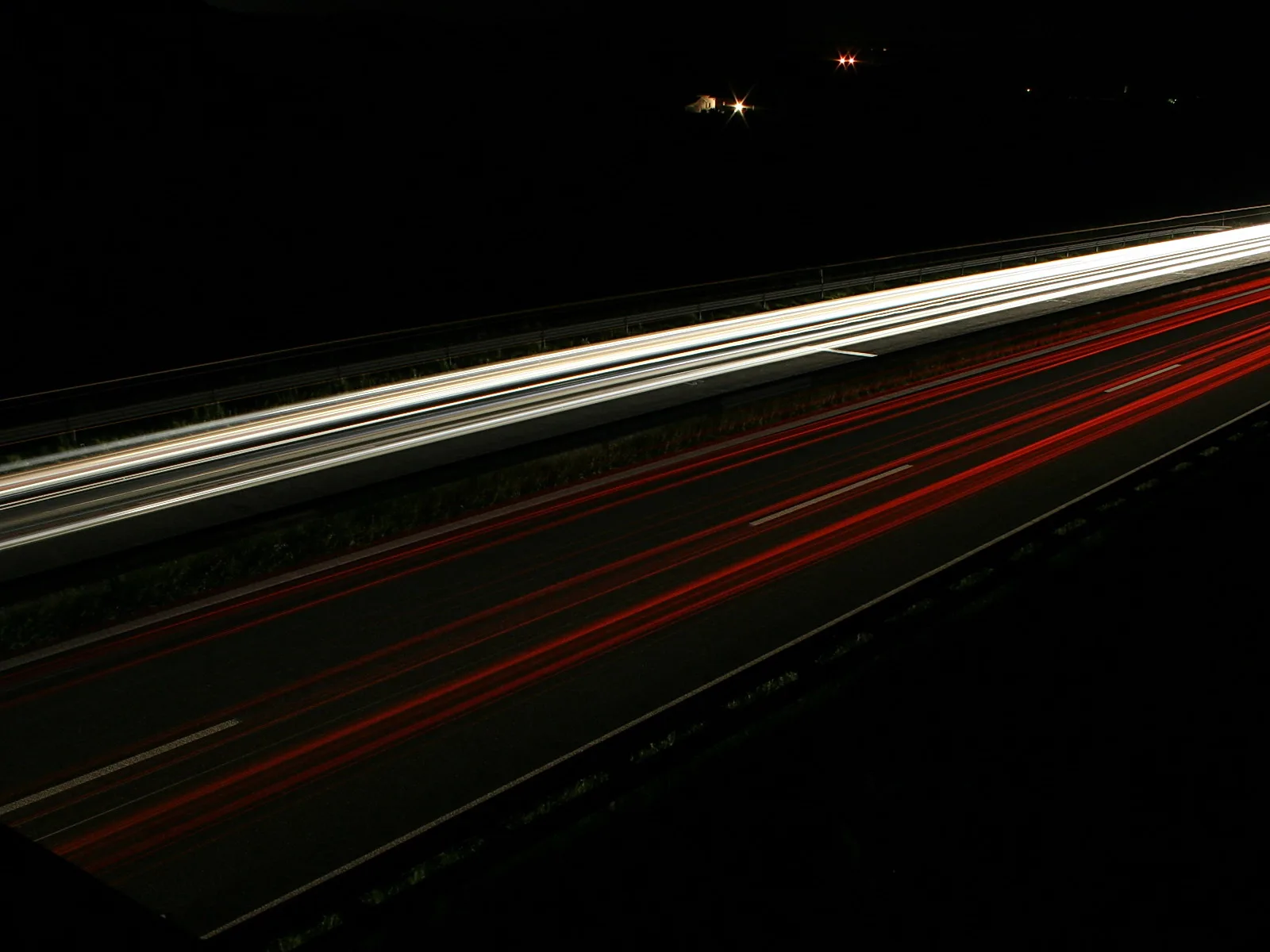 Langzeitbelichtung einer nächtlichen Autobahn mit weißen Lichtspuren in der oberen Hälfte und roten Rücklichtspuren auf den Fahrbahnen, dunkler Hintergrund.