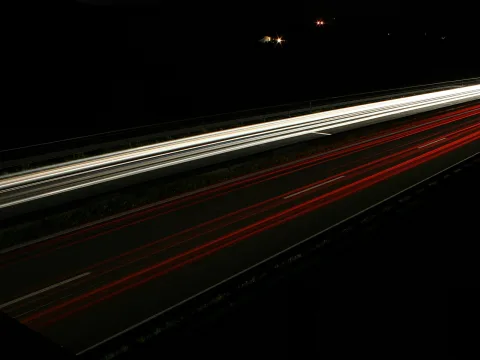 Langzeitbelichtung einer nächtlichen Autobahn mit weißen Lichtspuren in der oberen Hälfte und roten Rücklichtspuren auf den Fahrbahnen, dunkler Hintergrund.