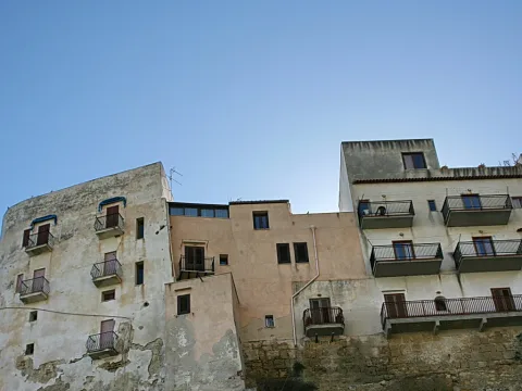Mehrstöckige, verwitterte Hausfassaden mit Balkonen und Fensterläden auf einer Felskante unter klarem blauem Himmel.