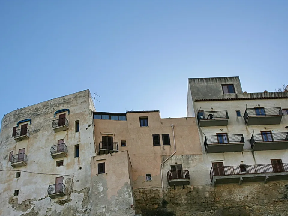 Mehrstöckige, verwitterte Hausfassaden mit Balkonen und Fensterläden auf einer Felskante unter klarem blauem Himmel.