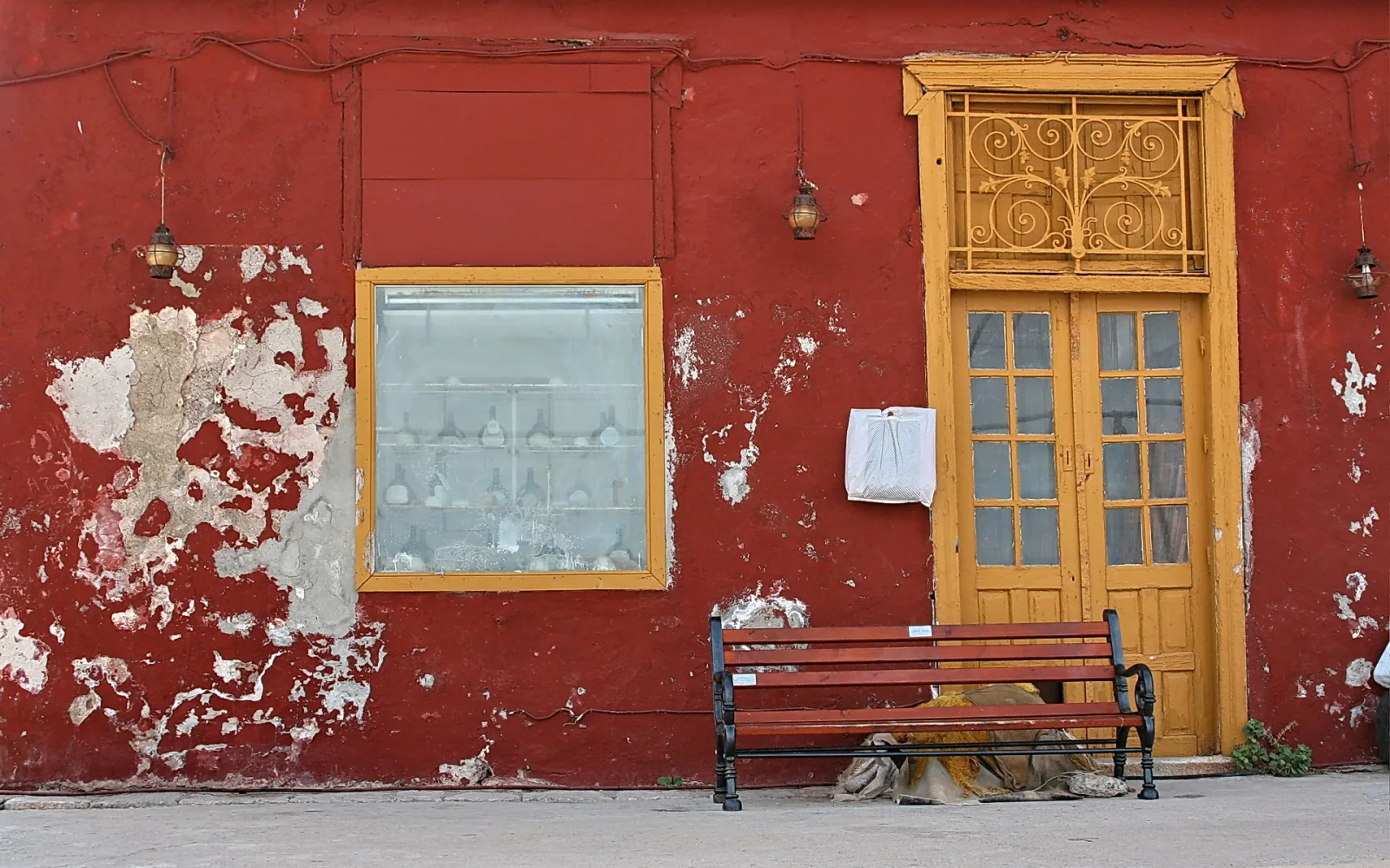 Rot verputzte Hauswand mit abblätternder Farbe, links ein Fenster mit gelbem Rahmen und Glasflaschen, rechts eine gelbe Doppeltür mit schmiedeeiserem Oberlicht, davor eine rotbraune Holzbank auf einem grauen Gehweg.