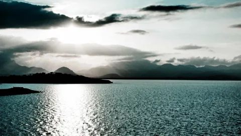 Ruhige Meeresbucht bei Sonnenuntergang mit silbrig glitzernder Wasseroberfläche, dunklen Hügeln und Inseln am Ufer, darüber dichte Wolken, durch die die Sonne scheint.