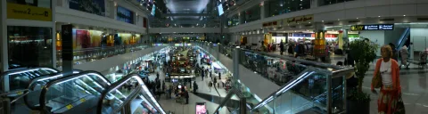 Große Flughafenhalle mit mehreren Ebenen, Rolltreppen im Vordergrund, Reisende auf dem Boden und Geschäften entlang der Galerien, links ein Schild mit Terminal 2.