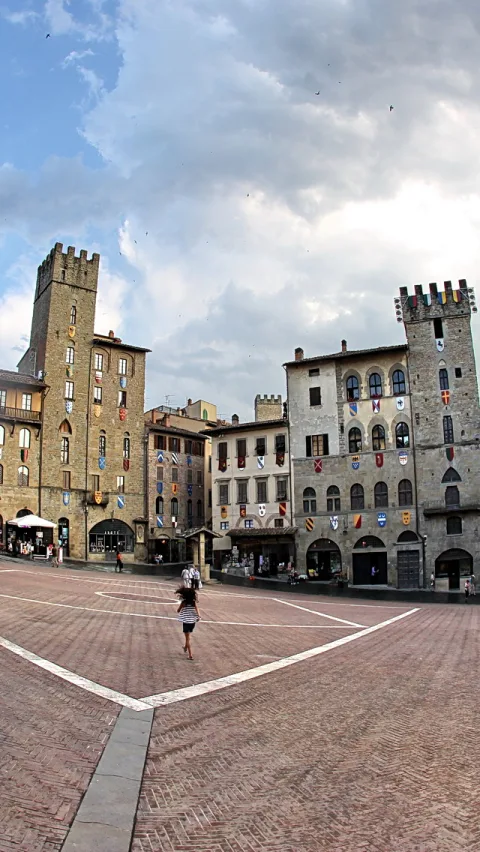 Gepflasterter Platz in Arezzo, umgeben von mittelalterlichen Gebäuden mit Türmen und Wappenfahnen, darüber ein bewölkter Himmel. Eine Frau geht über den Platz.