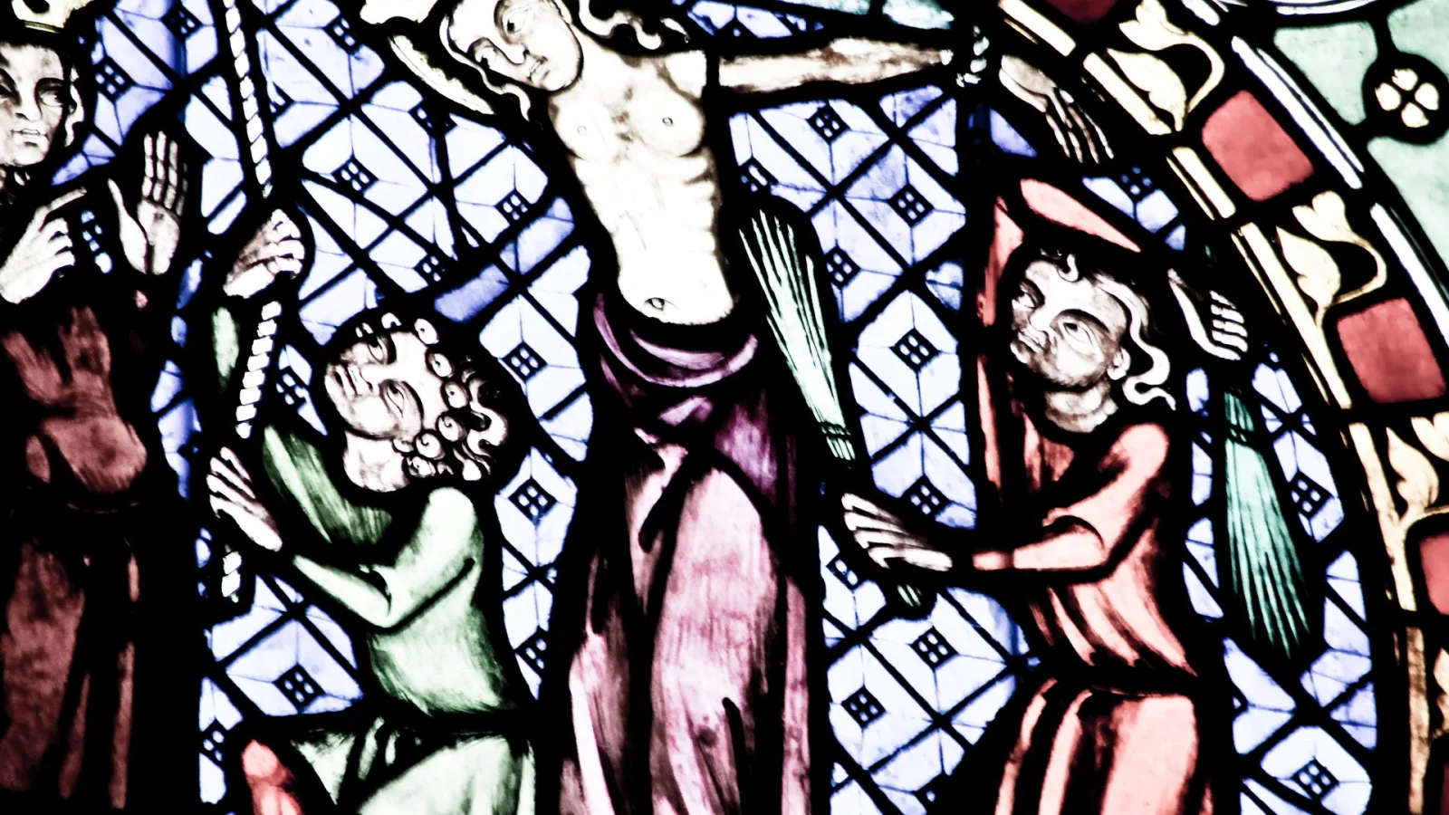Buntglasfenster mit einer Kreuzigungsszene: Christus am Kreuz in der Mitte, flankiert von zwei Figuren in grünen und roten Gewändern, vor einem blau-weißen Rautenmuster.
