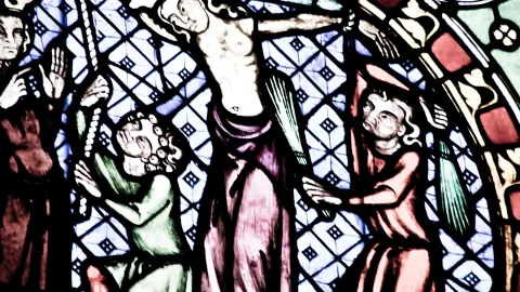 Buntglasfenster mit einer Kreuzigungsszene: Christus am Kreuz in der Mitte, flankiert von zwei Figuren in grünen und roten Gewändern, vor einem blau-weißen Rautenmuster.