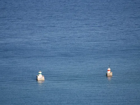 Zwei kleine Hafenfeuer mit Betonsockel auf blauem, ruhigem Meer; links ein weiß-grüner, rechts ein rot-weißer Turm.
