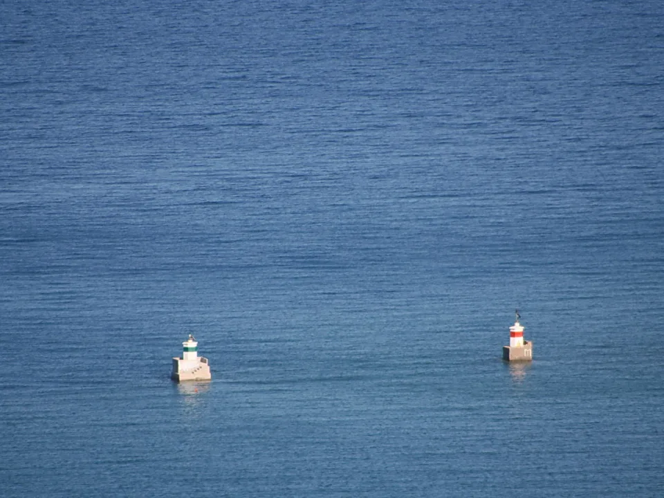 Zwei kleine Hafenfeuer mit Betonsockel auf blauem, ruhigem Meer; links ein weiß-grüner, rechts ein rot-weißer Turm.