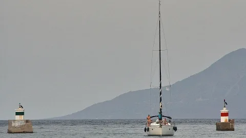 Ein weißes Segelboot fährt auf ein Hafenbecken zu, flankiert von zwei kleinen Molen mit Leuchttürmen, dahinter eine bergige Küste im grauen Dunst.