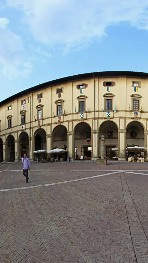 Gebogene gelbe Fassade mit Rundbogenarkaden auf einem gepflasterten Platz, davor Sonnenschirme und Tische eines Cafés, links läuft ein Mann über den Platz.