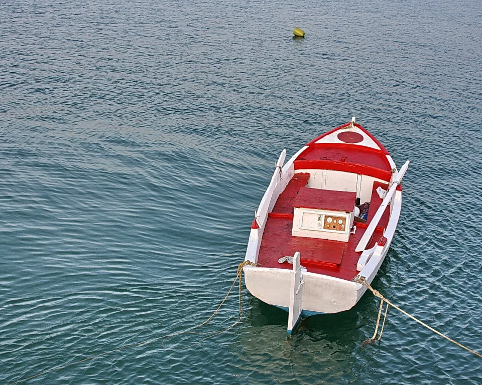 Ein rot-weiß lackiertes Holzboot liegt vertäut auf einer ruhigen, blaugrünen Wasserfläche, im Hintergrund schwimmt eine gelbe Boje.
