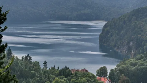 Ein langgestreckter Bergsee unter bewaldeten Hängen, im Vordergrund Bäume und zwei rotgedeckte Häuser am Ufer.