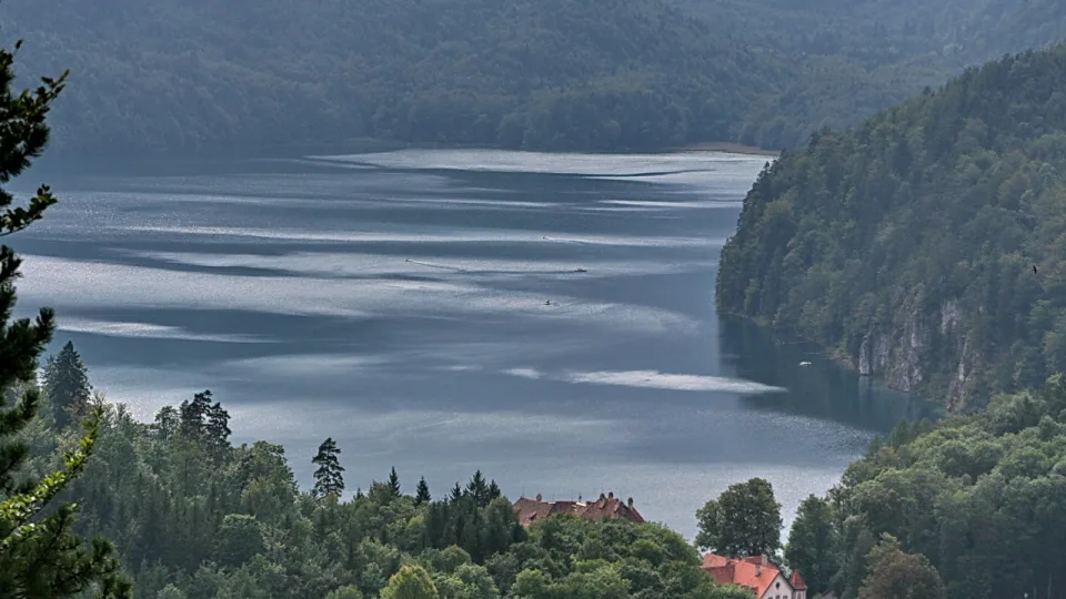 Ein langgestreckter Bergsee unter bewaldeten Hängen, im Vordergrund Bäume und zwei rotgedeckte Häuser am Ufer.