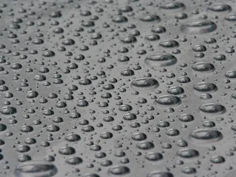 Viele Wassertropfen unterschiedlicher Größe liegen auf einer glatten grauen Oberfläche, die Tropfen spiegeln das Licht.