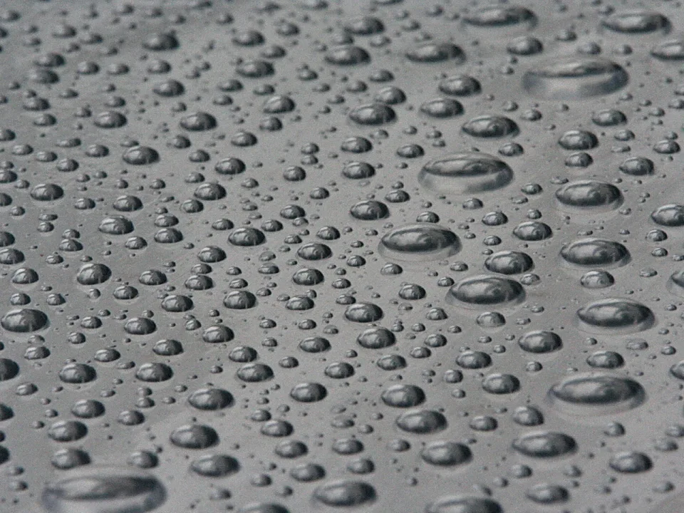 Viele Wassertropfen unterschiedlicher Größe liegen auf einer glatten grauen Oberfläche, die Tropfen spiegeln das Licht.