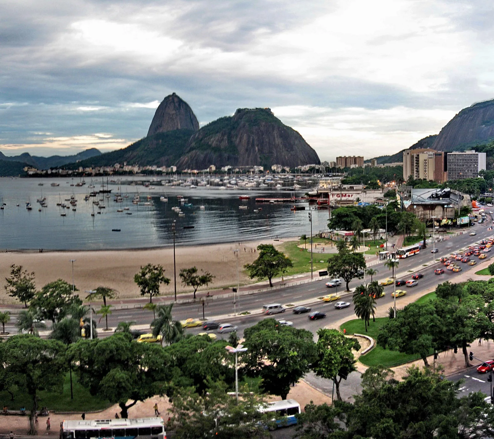 Blick über die Bucht von Botafogo in Rio de Janeiro auf den Zuckerhut und einen Berg mit Bebauung, davor Boote im Wasser, ein Sandstrand, eine mehrspurige Straße mit Autos, Bussen und gelben Taxis sowie Bäume und Hochhäuser, darüber bewölkter Himmel.