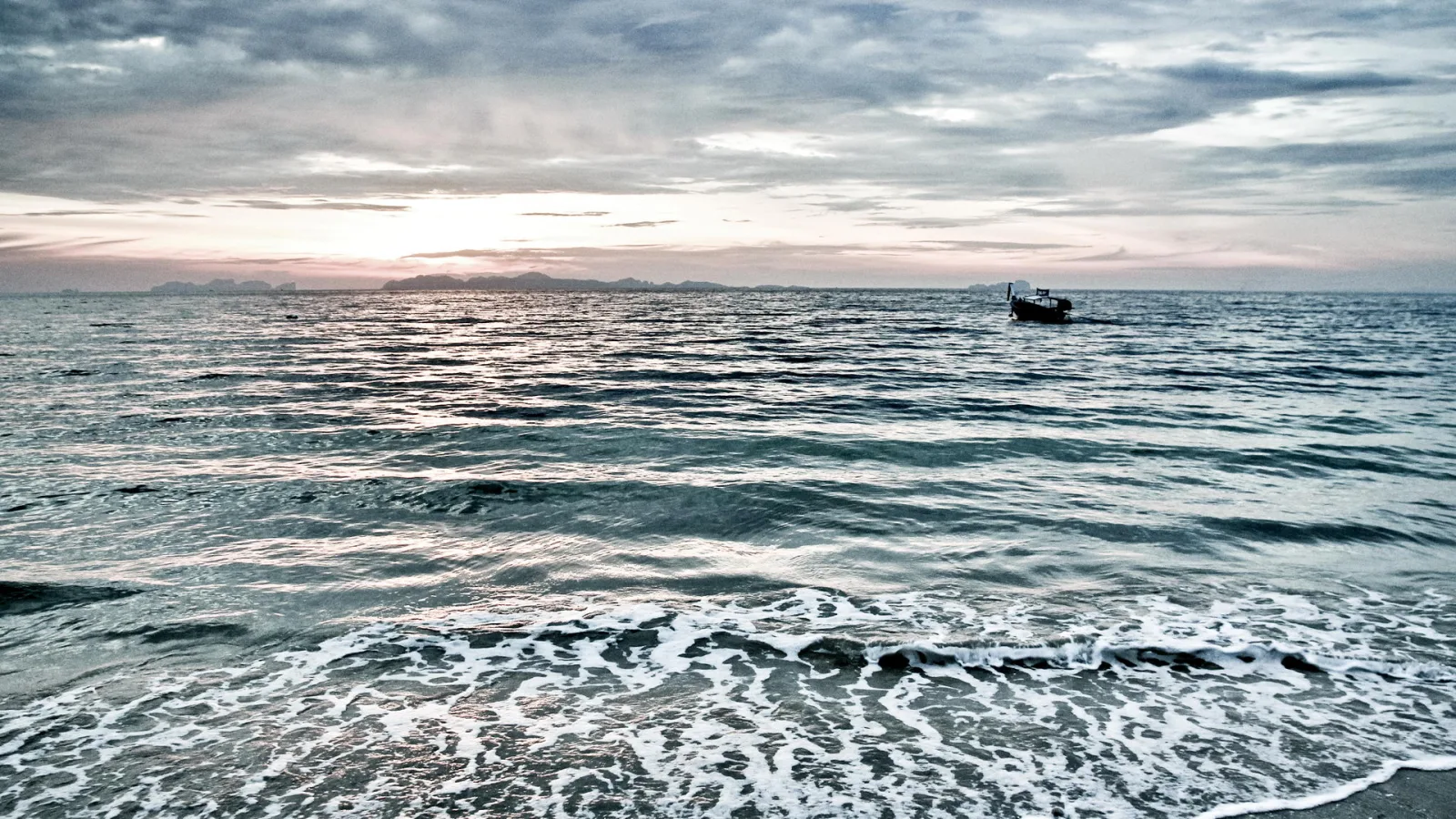 Ruhiges Meer unter bewölktem Abendhimmel, im Hintergrund flache Inseln am Horizont, rechts ein kleines Boot und vorne Brandung mit Schaum am Strand.