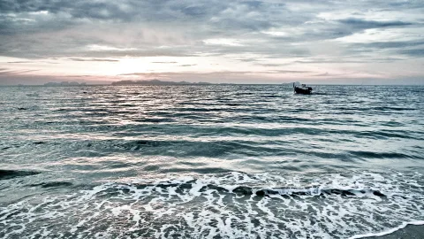 Ruhiges Meer unter bewölktem Abendhimmel, im Hintergrund flache Inseln am Horizont, rechts ein kleines Boot und vorne Brandung mit Schaum am Strand.