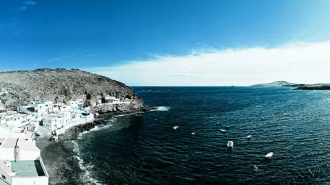 Weiße Häuser auf einem Felsvorsprung über dunklem Meer, davor kleine Boote auf den Wellen und eine flache Insel am Horizont.