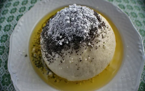 Ein helles Germknödel mit dunklen Mohnsamen und Puderzucker oben, liegt auf goldener Flüssigkeit auf einem weißen Teller.