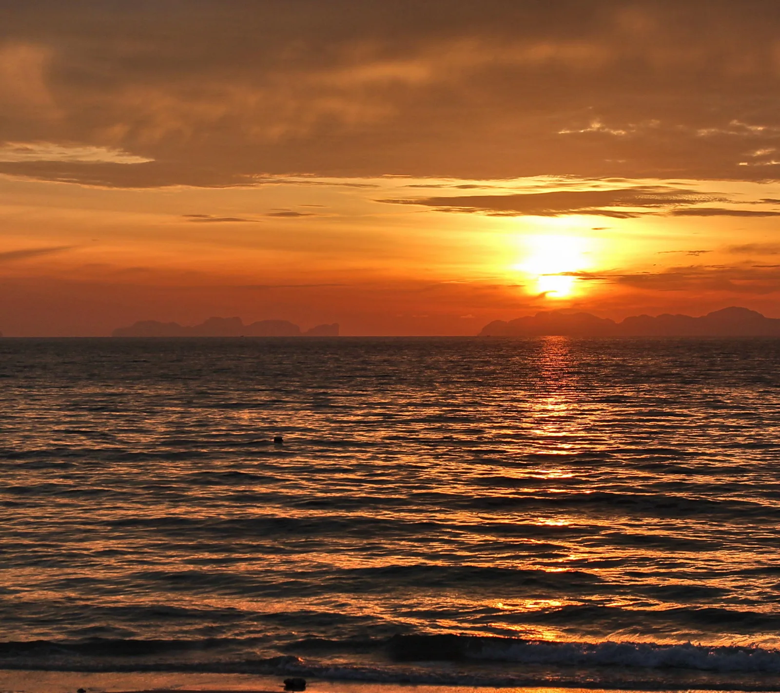Sonnenuntergang über dem Meer, die Sonne sinkt hinter einem orangefarbenen Wolkenband. Am Horizont zeichnen sich dunkle Inselsilhouetten ab, im Vordergrund glitzert die Wasseroberfläche.