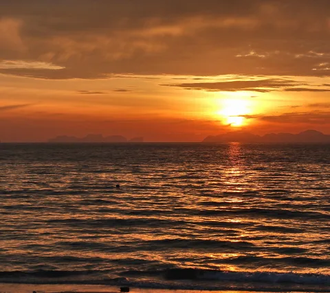 Sonnenuntergang über dem Meer, die Sonne sinkt hinter einem orangefarbenen Wolkenband. Am Horizont zeichnen sich dunkle Inselsilhouetten ab, im Vordergrund glitzert die Wasseroberfläche.