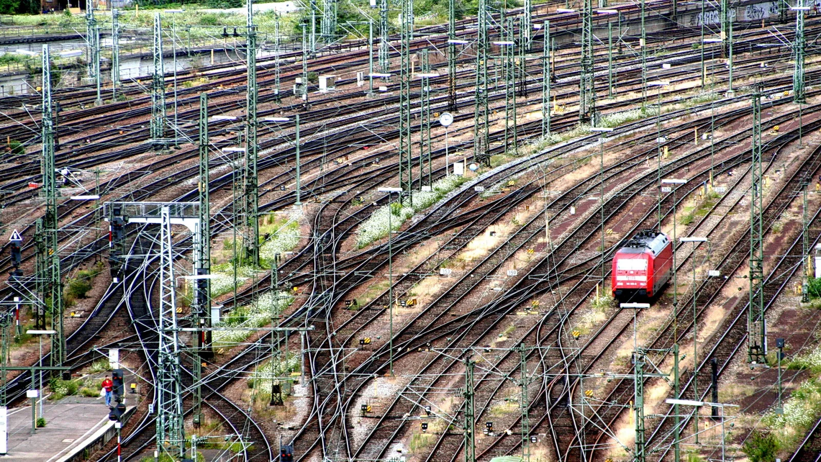Viele Gleise und Oberleitungsmasten auf einem Bahngelände, rechts fährt eine rote Lokomotive durch die Weichen.