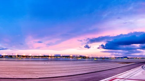 Leere Landebahn eines Flughafens bei Sonnenuntergang, am Horizont leuchten Lichter, darüber ein violett-rosa Himmel mit dunklen Wolken.