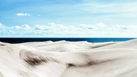 Helle, wellige Sanddünen im Vordergrund, dahinter dunkelblaues Meer unter einem leicht bewölkten, hellblauen Himmel.
