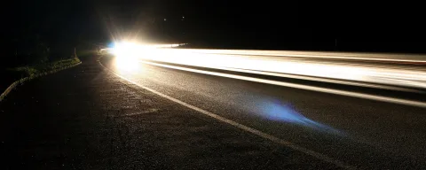 Langzeitbelichtung einer nächtlichen Straße mit Scheinwerferlichtern, die als helle Streifen nach rechts verlaufen, und einem Seitenstreifen links im Bild.