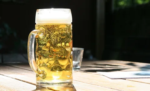 Ein Glas Helles mit Schaumkrone steht auf einem Holztisch im Sonnenlicht, im Hintergrund ein leeres Glas vor dunklem Grund.