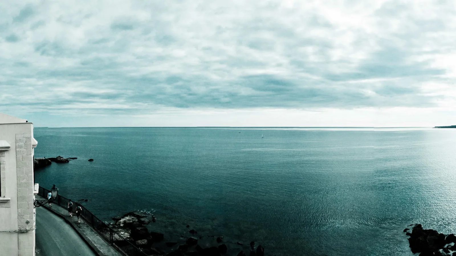Weite Meeresfläche unter bewölktem Himmel, links ein weißes Gebäude mit Geländer und Straße am Ufer, im Vordergrund dunkle Felsen, am Horizont ein kleines Segelboot.
