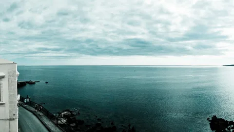 Weite Meeresfläche unter bewölktem Himmel, links ein weißes Gebäude mit Geländer und Straße am Ufer, im Vordergrund dunkle Felsen, am Horizont ein kleines Segelboot.