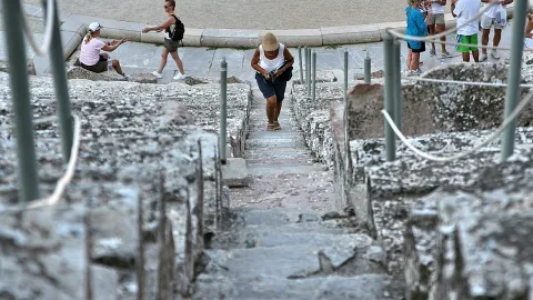 Ein Mann mit Hut und weißem Trägershirt steigt eine schmale Steintreppe zwischen alten Mauerresten hinauf, im Hintergrund sitzen und gehen weitere Besucher.