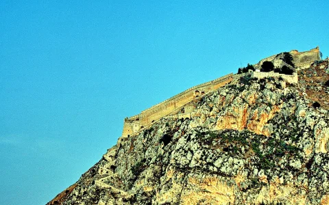 Alte Festungsmauer mit Türmen und Treppen auf einem steilen, felsigen Berggipfel vor klarem blauem Himmel.