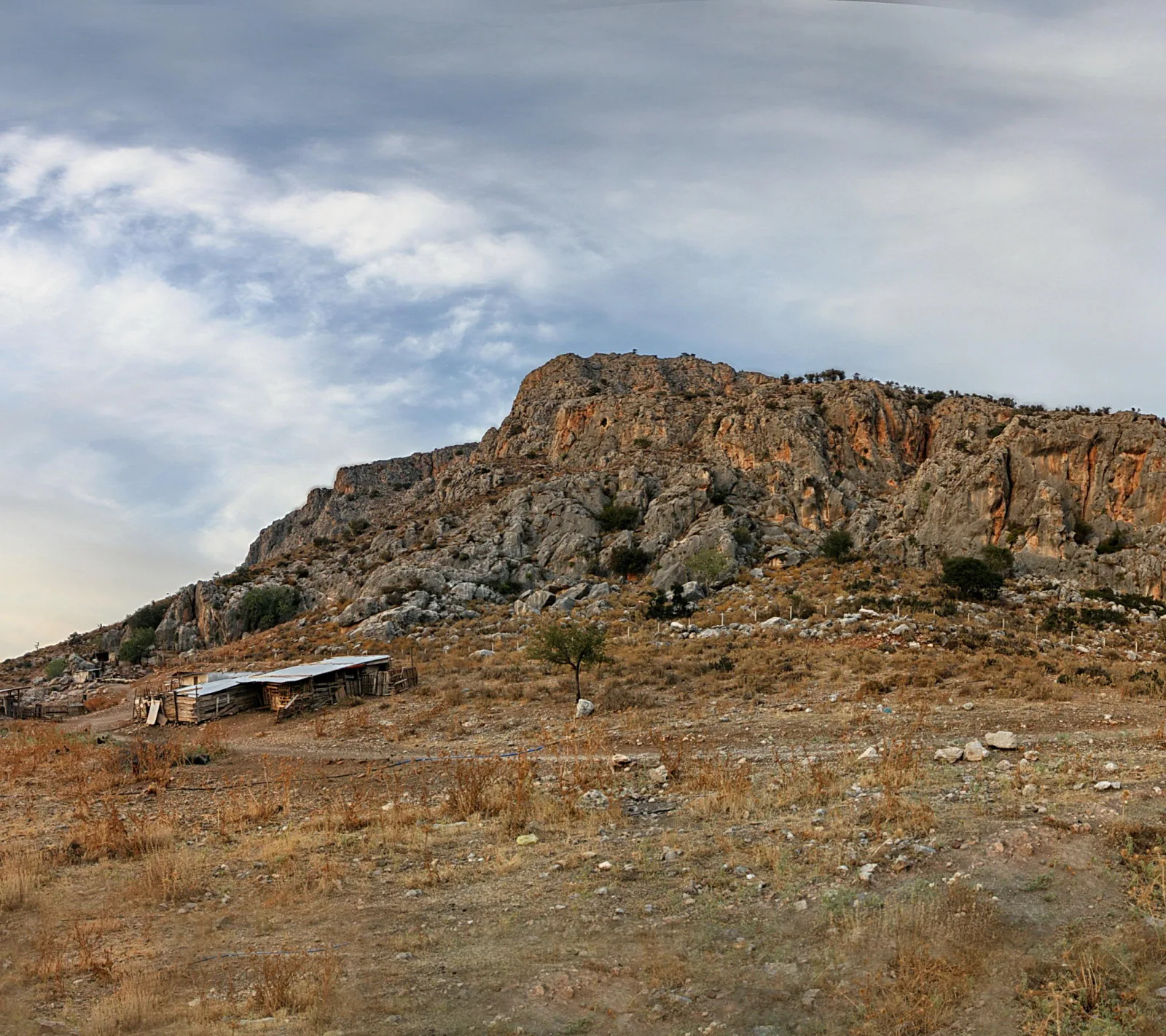 Felsiger Berg mit orangefarbenen Felswänden, davor trockenes Gras, ein einzelner Baum und eine kleine Holzhütte mit Wellblechdach unter bewölktem Himmel.