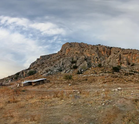 Felsiger Berg mit orangefarbenen Felswänden, davor trockenes Gras, ein einzelner Baum und eine kleine Holzhütte mit Wellblechdach unter bewölktem Himmel.