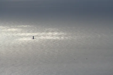 Kleiner dunkler Leuchtturm auf einer glitzernden, silbrig grauen Meeresfläche unter diesigem Himmel.