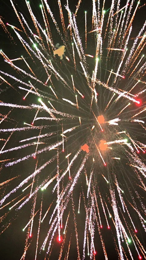 Feuerwerk mit vielen weißen und goldenen Funkenbahnen, die von einem Zentrum nach außen strahlen, dazwischen einzelne rote und orangefarbene Explosionen vor dunklem Nachthimmel.