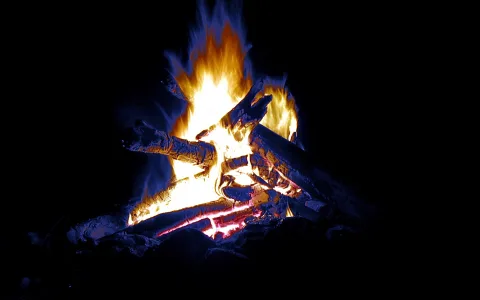 Lagerfeuer in völliger Dunkelheit, Holzscheite liegen in den Flammen, die gelb und blau lodern, an den Scheiten glüht rotes Holz.