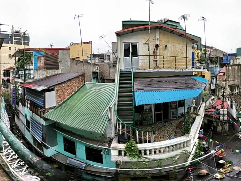 Fischaugen-Ansicht eines dicht bebauten Hinterhofs in Hanoi mit mehrstöckigen Häusern, grünen und blauen Wellblechdächern, einer Außentreppe und einem kleinen Straßenmarkt mit Körben und Schalen am Boden.