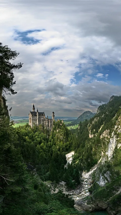 Schloss Neuschwanstein auf einem bewaldeten Felsen über einer Schlucht mit Wildbach, darüber bewölkter Himmel.