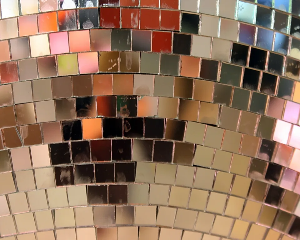 Nahaufnahme einer Discokugel aus kleinen, quadratischen Spiegelkacheln in Gold-, Rot- und Brauntönen, in leicht gebogenen Reihen angeordnet.