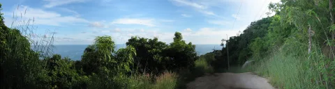 Schotterweg führt bergab durch dichtes grünes Gebüsch, im Hintergrund blaues Meer unter einem leicht bewölkten Himmel, rechts ein Strommast.