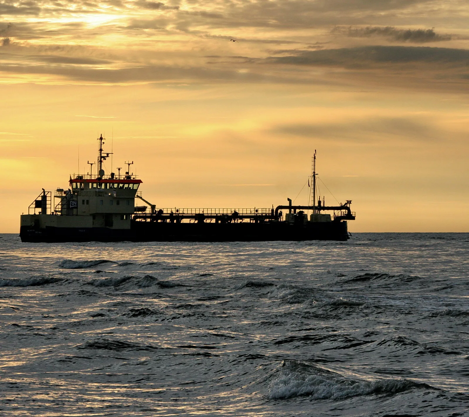 Ein dunkles Frachtschiff mit weißem Aufbau und roter Reling fährt bei Sonnenuntergang über bewegte Ostsee-Wellen.