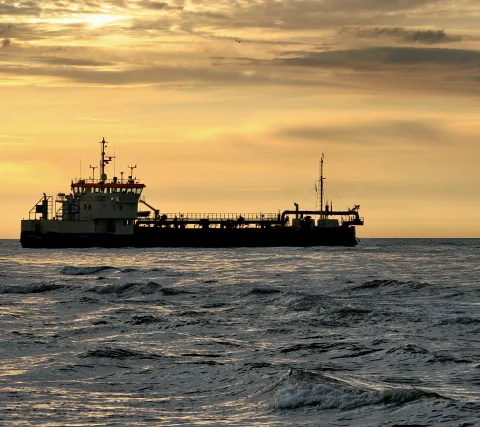 Ein dunkles Frachtschiff mit weißem Aufbau und roter Reling fährt bei Sonnenuntergang über bewegte Ostsee-Wellen.
