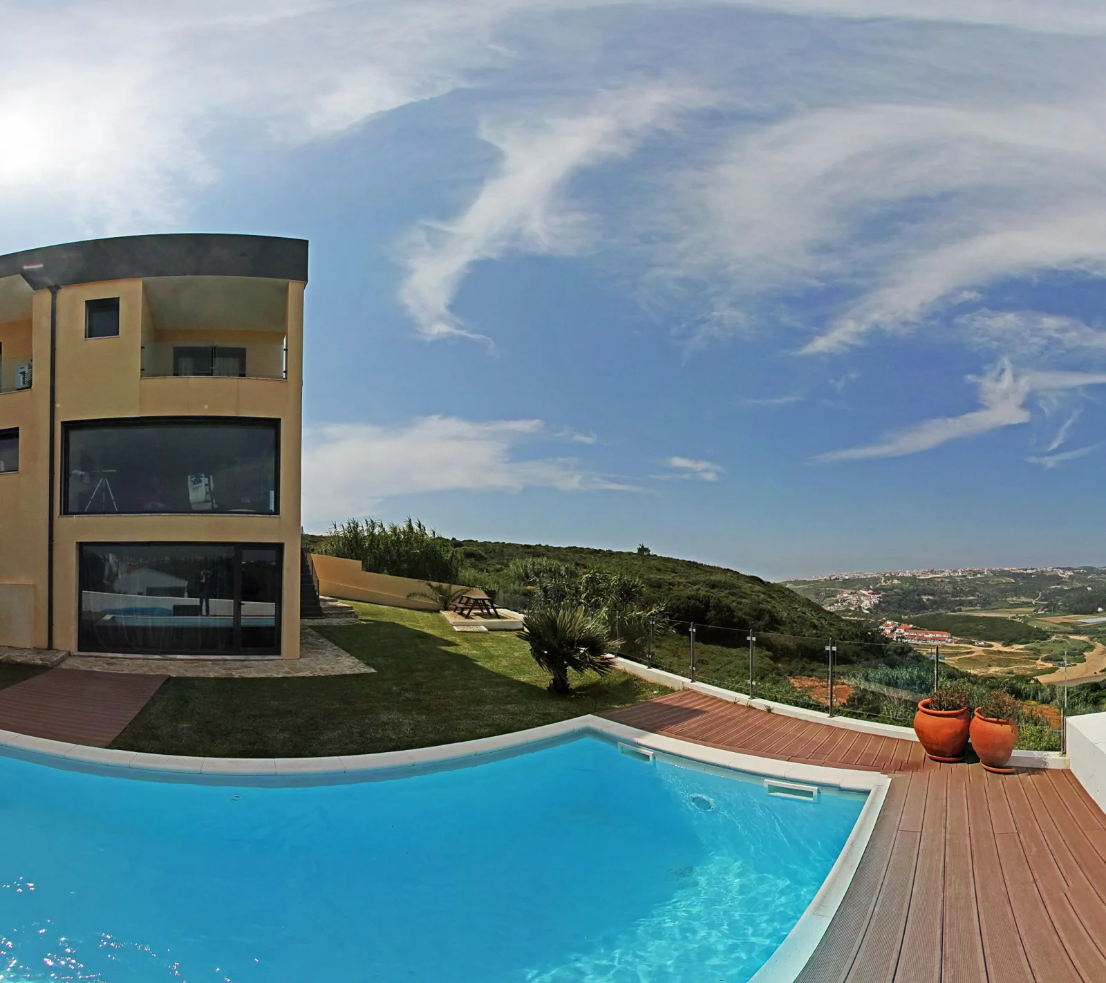 Gelbe Villa mit großen Glasfronten, davor ein türkisblauer Pool auf einer Holzterrasse mit Terrakottatöpfen, im Hintergrund Hügel und ein blauer Himmel mit Schleierwolken.