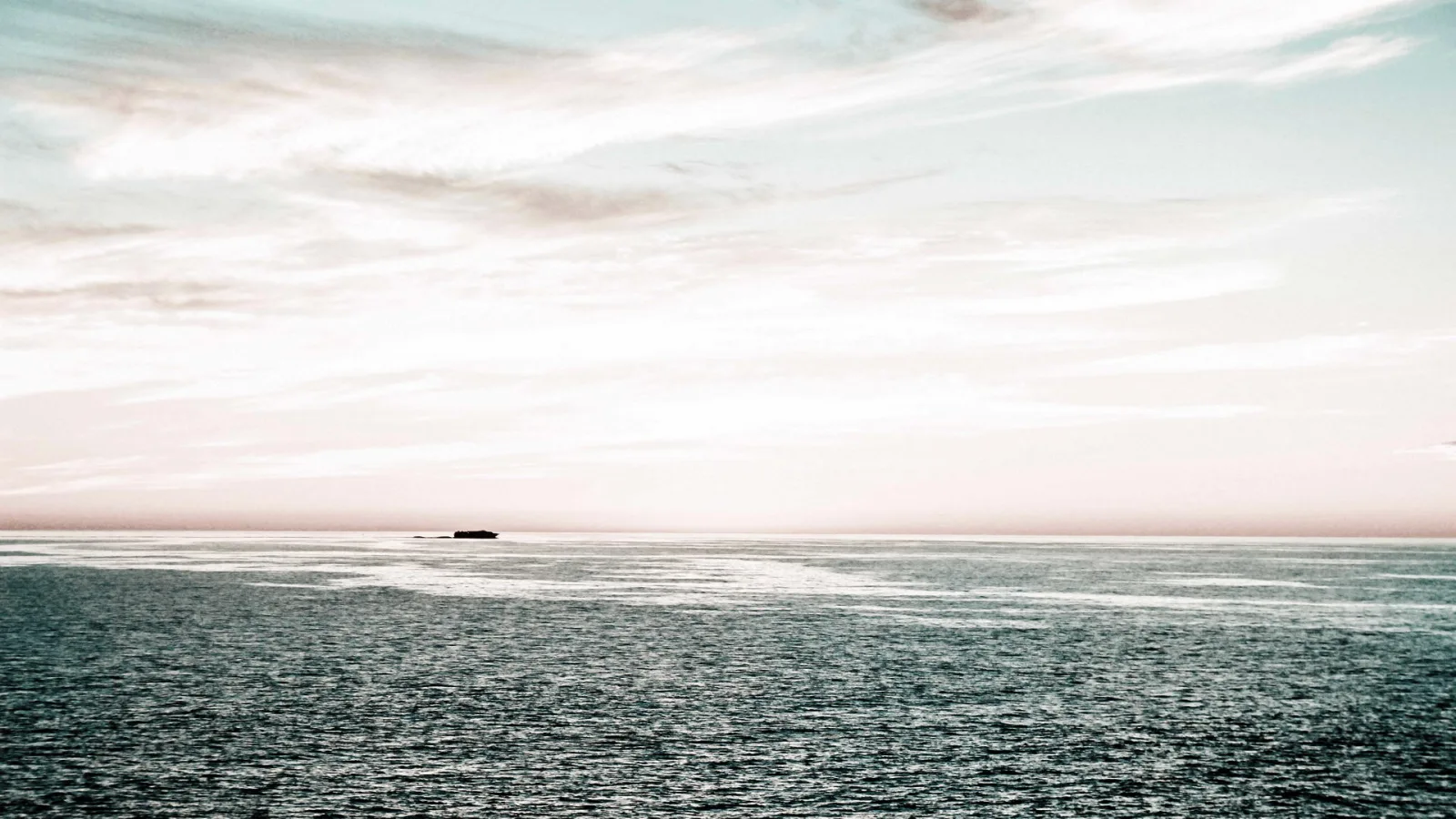 Ruhige Meeresfläche unter einem hellen, rosa getönten Himmel mit Schleierwolken, am Horizont eine kleine dunkle Insel.