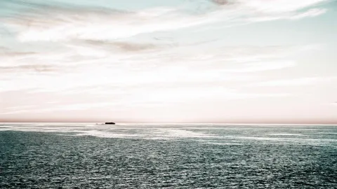 Ruhige Meeresfläche unter einem hellen, rosa getönten Himmel mit Schleierwolken, am Horizont eine kleine dunkle Insel.