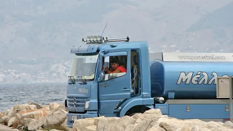Blauer Tankwagen mit Aufschrift Μέλλος steht auf Felsbrocken am Meer, ein Fahrer sitzt in der Kabine, im Hintergrund hügelige Küste im Dunst.